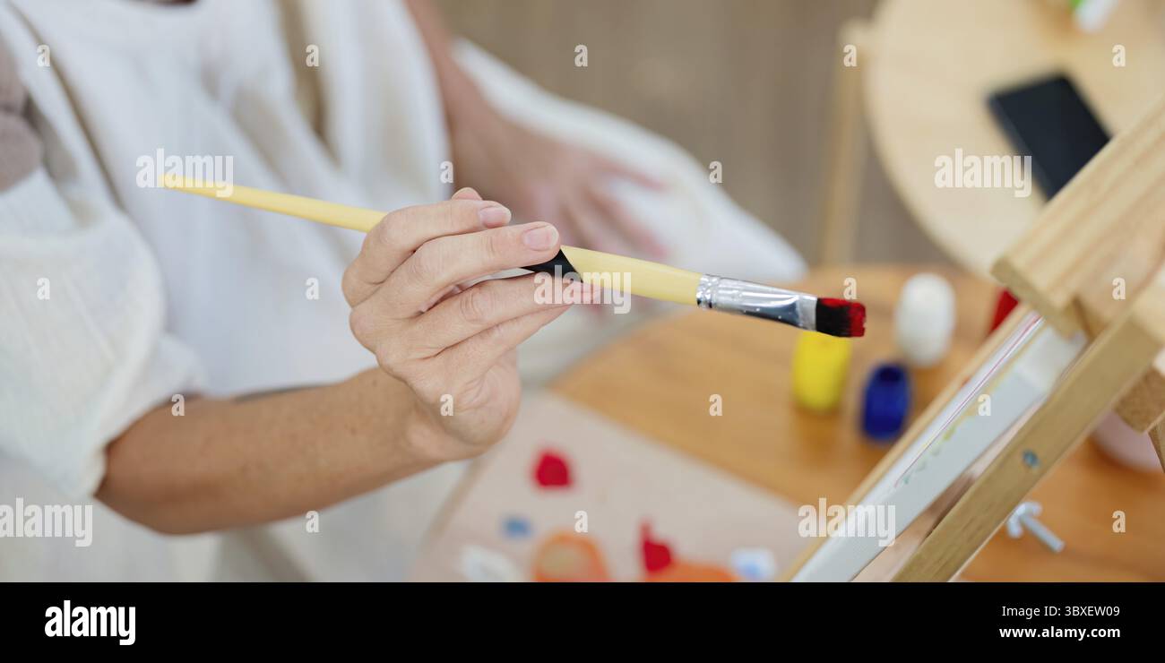 Vista dettagliata di una mano anziana che tiene delicatamente un pennello sopra una tavolozza di colori vivaci, preparandosi a creare arte Foto Stock