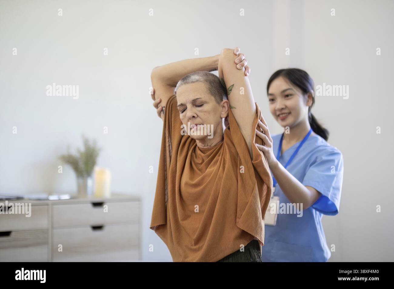Infermiera asiatica che assiste una donna anziana con esercizi di allungamento del braccio a casa, concentrandosi sulla cura degli anziani e sulla terapia fisica Foto Stock