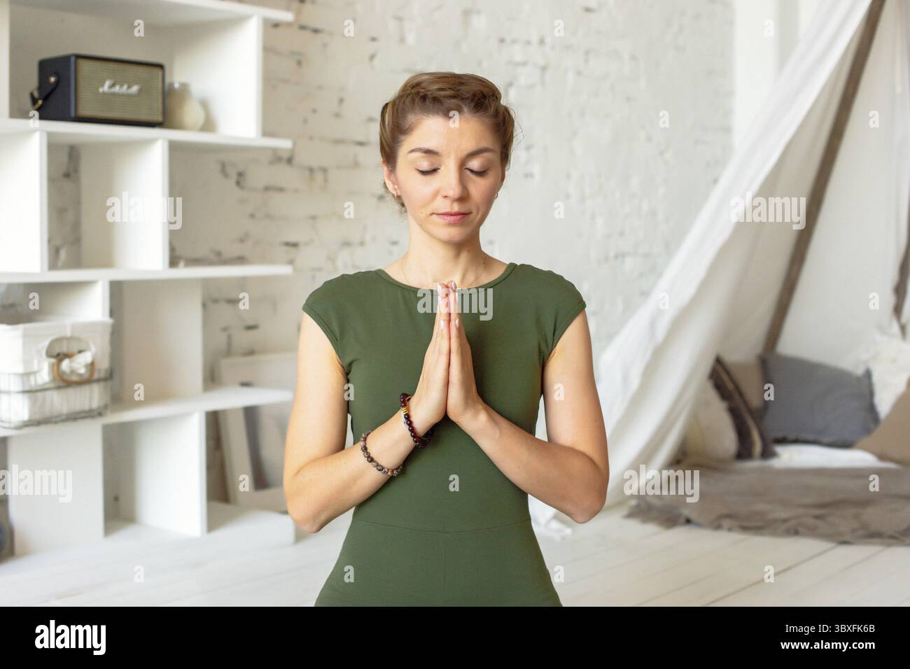 Bella donna slanciata in una maglietta verde, seduta in posa yoga, in una stanza luminosa, con i palmi piegati insieme al petto e chiudendo gli occhi Foto Stock