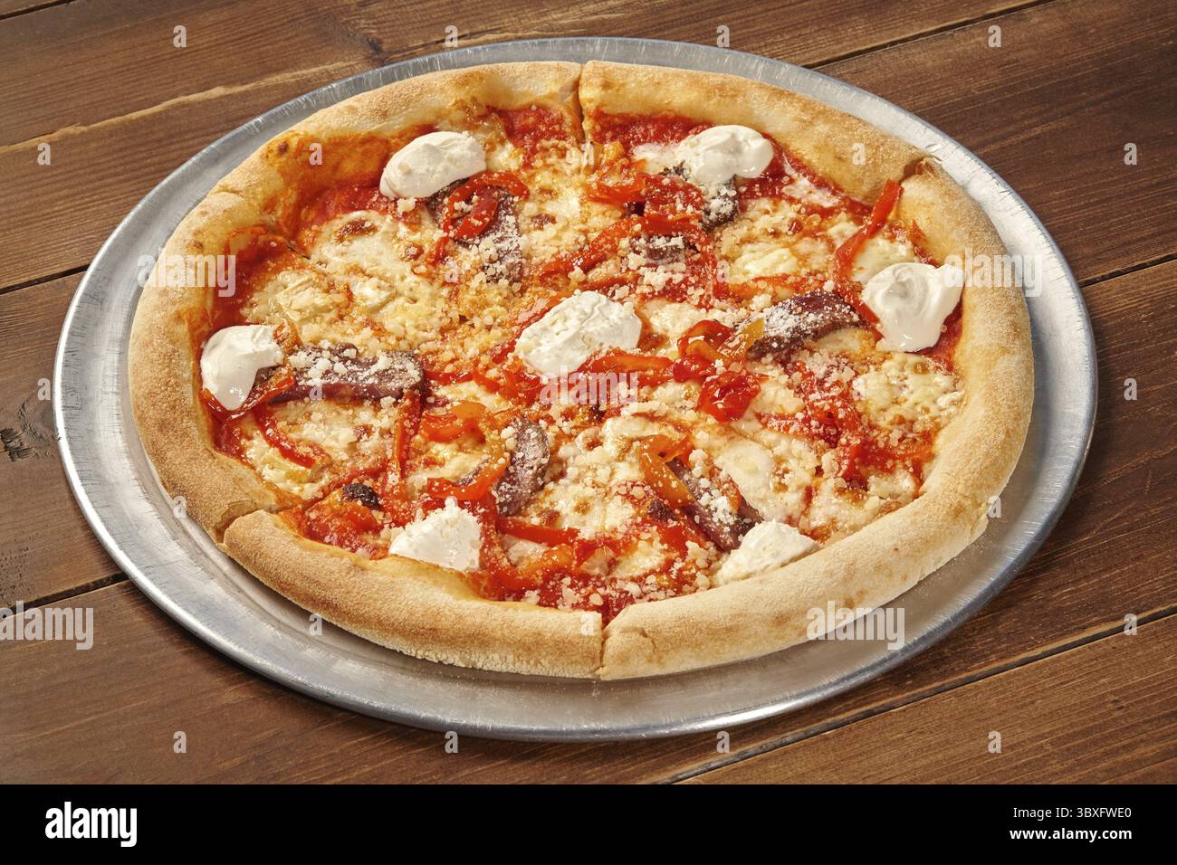 La pizza rotonda con acciughe, peperoni rossi, salsa cremosa e formaggio grattugiato si trova su un vassoio di metallo su un tavolo di legno Foto Stock