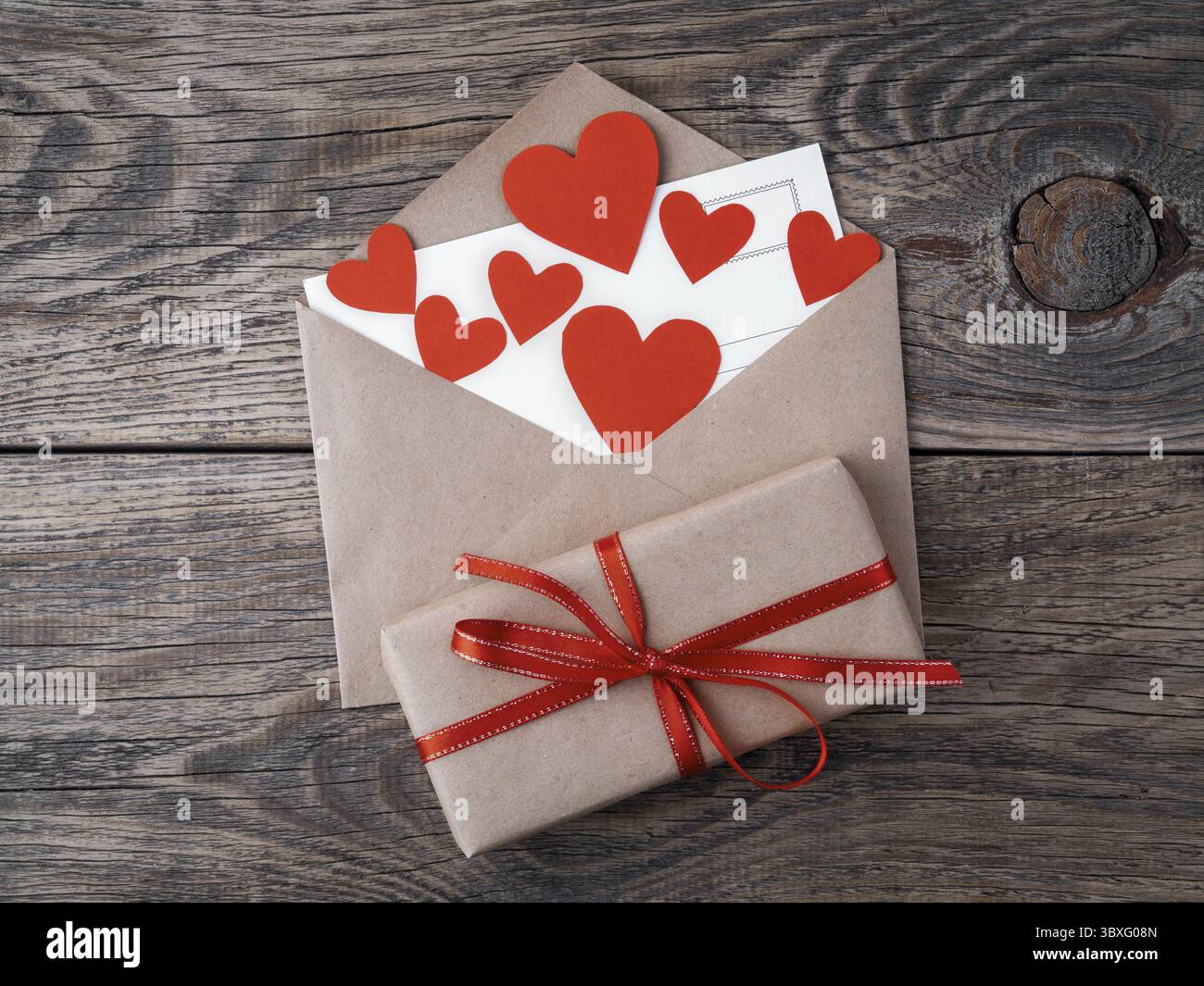 Karte und rote Herzen im offenen Umschlag aus braunem Kraftpapier. Geschenkkasten mit rotem Band auf hoelzernen alten Jahrgang Hintergrund. Gruesse An Foto Stock