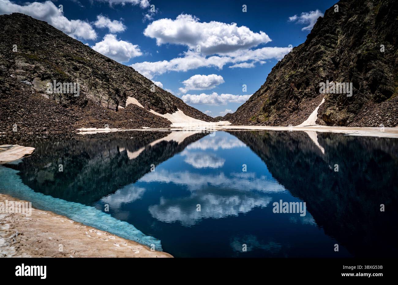 Un tranquillo lago di montagna che riflette il cielo e le scogliere rocciose circostanti. L'acqua è calma, con un cielo blu limpido e soffici nuvole. La l Foto Stock