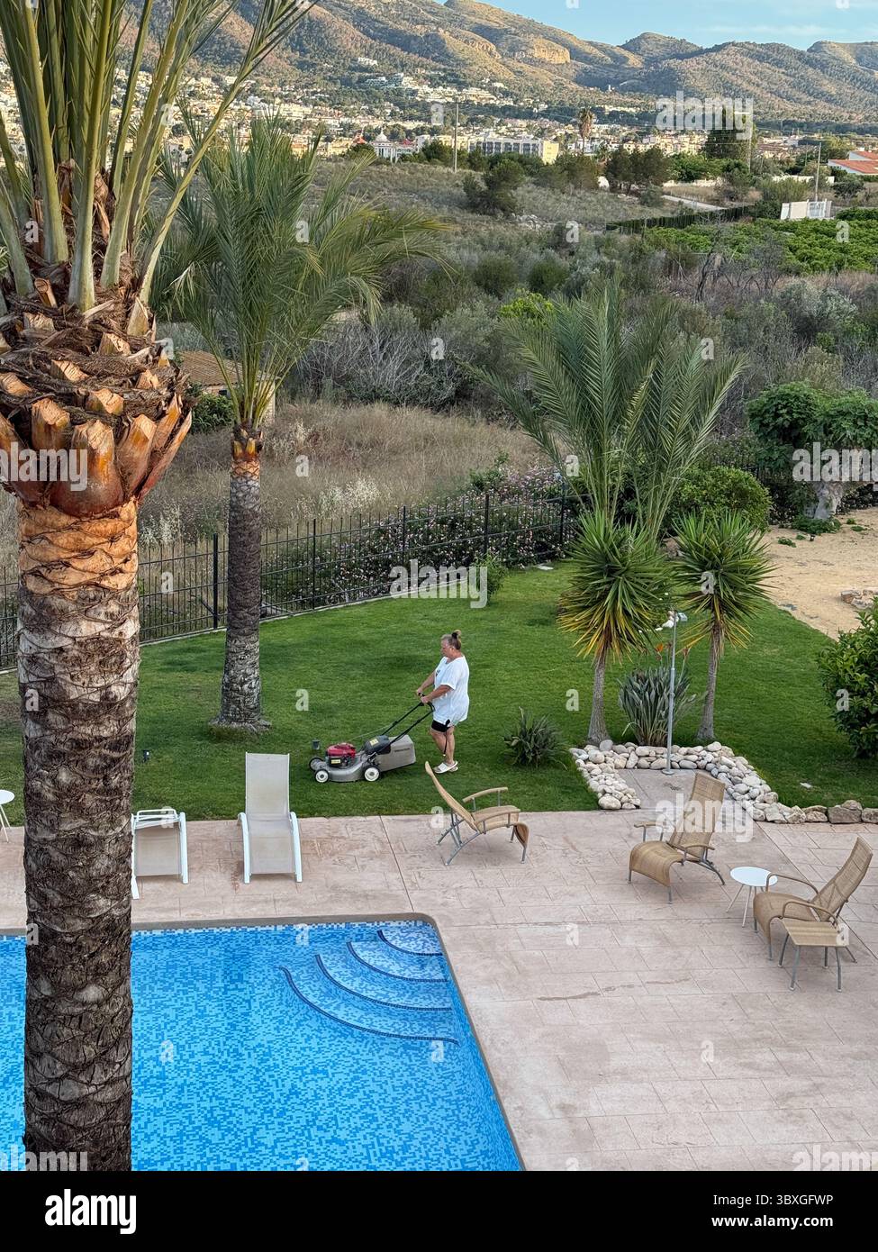 La persona sta falciando un vivace prato verde accanto ad una piscina limpida, con palme e montagne lontane che esaltano il tranquillo ambiente esterno Foto Stock