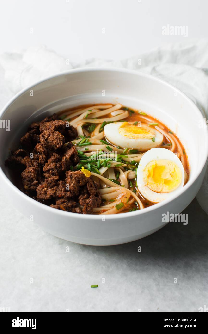 Vista dall'alto della zuppa piccante di spaghetti con uova e pezzi di manzo in una ciotola bianca, vista dall'alto delle uova di spaghetti in un brodo piccante con scalpelli Foto Stock