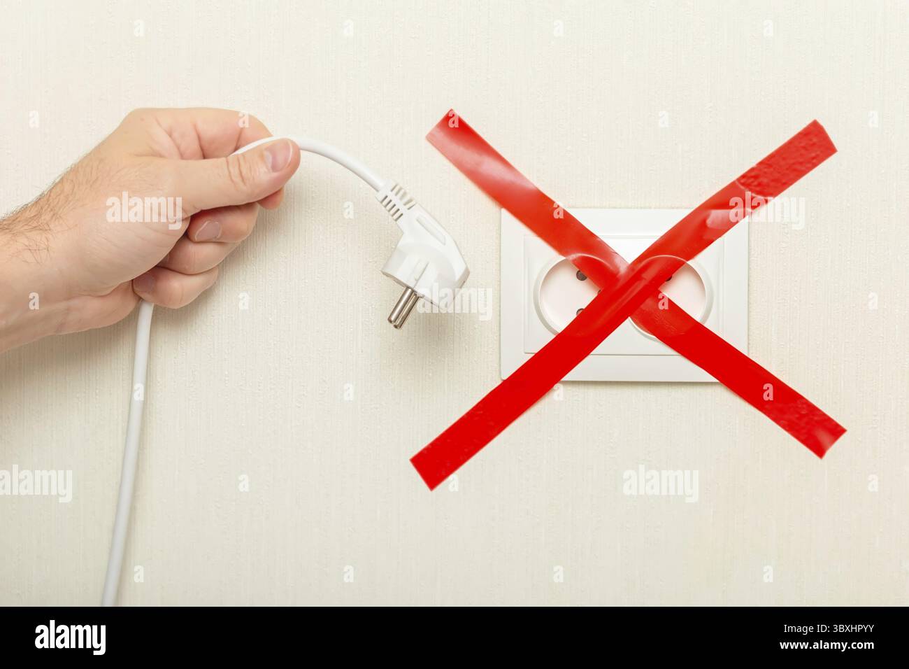 Una mano mans tiene un cavo bianco cadente con una spina vicino a una presa elettrica, legata con un nastro rosso trasversale. Il concetto di crisi energetica in E. Foto Stock