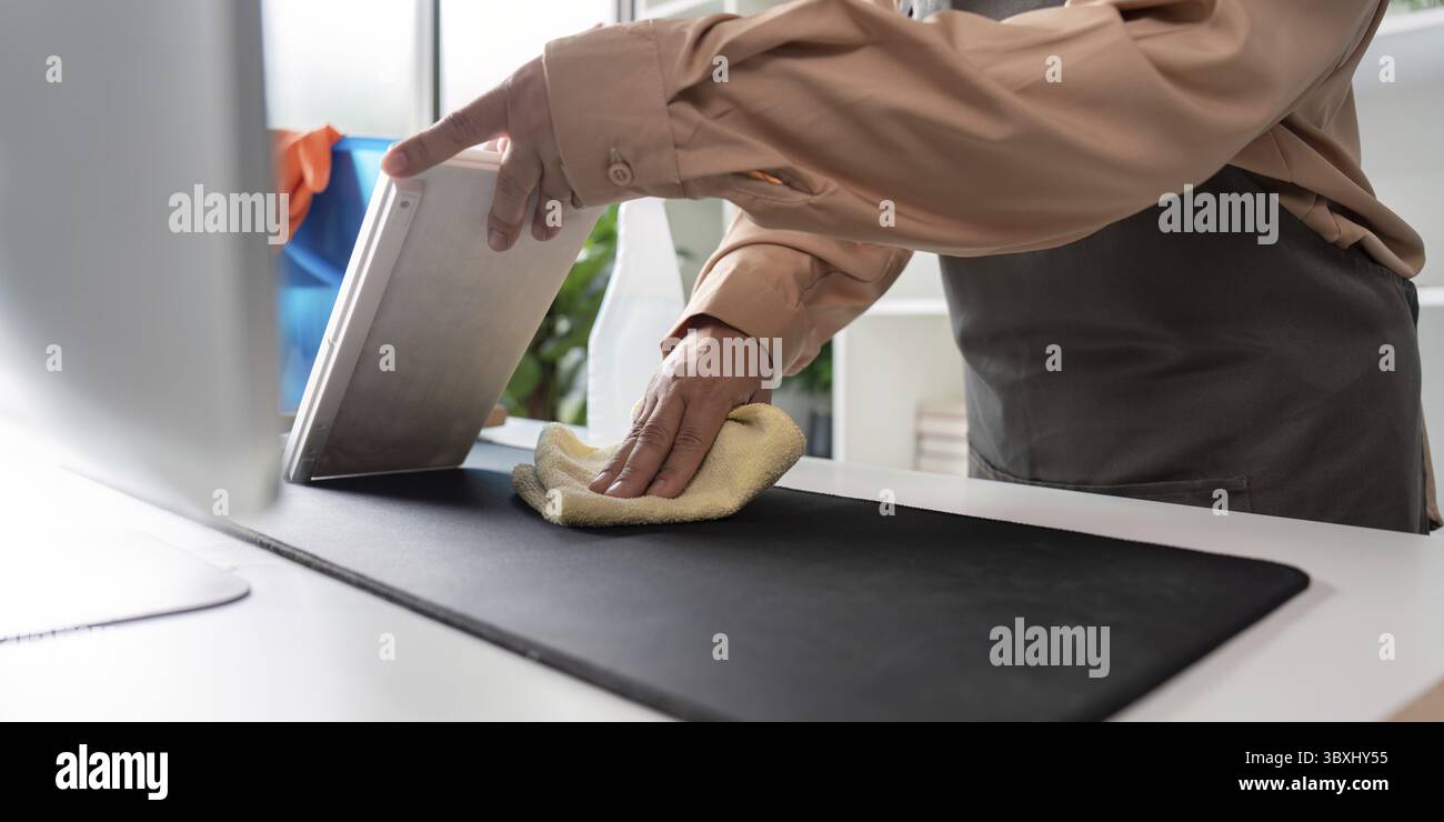 Una cameriera professionale che pulisce una moderna scrivania con un panno e un tablet in una camera luminosa e ben illuminata Foto Stock