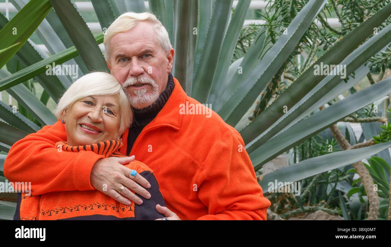 Felice anziani bella coppia sposata con i capelli grigi in arancione sportswear contro uno sfondo di serra piante esotiche Foto Stock