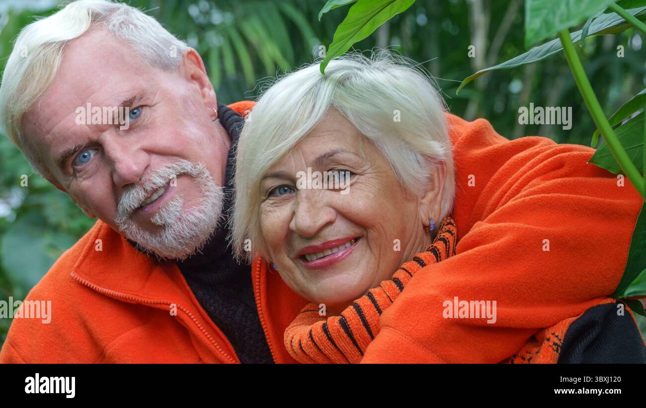 Felice anziani bella coppia sposata con i capelli grigi in arancione sportswear contro uno sfondo di serra piante esotiche Foto Stock