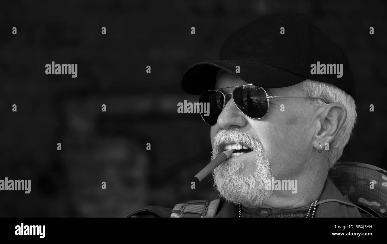 Ritratto di fiducioso uomo anziano indossare abbigliamento protettivo in occhiali da sole con un sigaro contro lo sfondo di vecchi muri di cemento Foto Stock