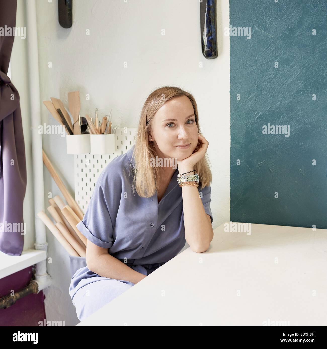 Donna d'affari seduta al tavolo e sorridente in un laboratorio di ceramica. Giovane donna d'affari sul posto di lavoro in ceramica. Allegra giovane donna in abito blu Foto Stock
