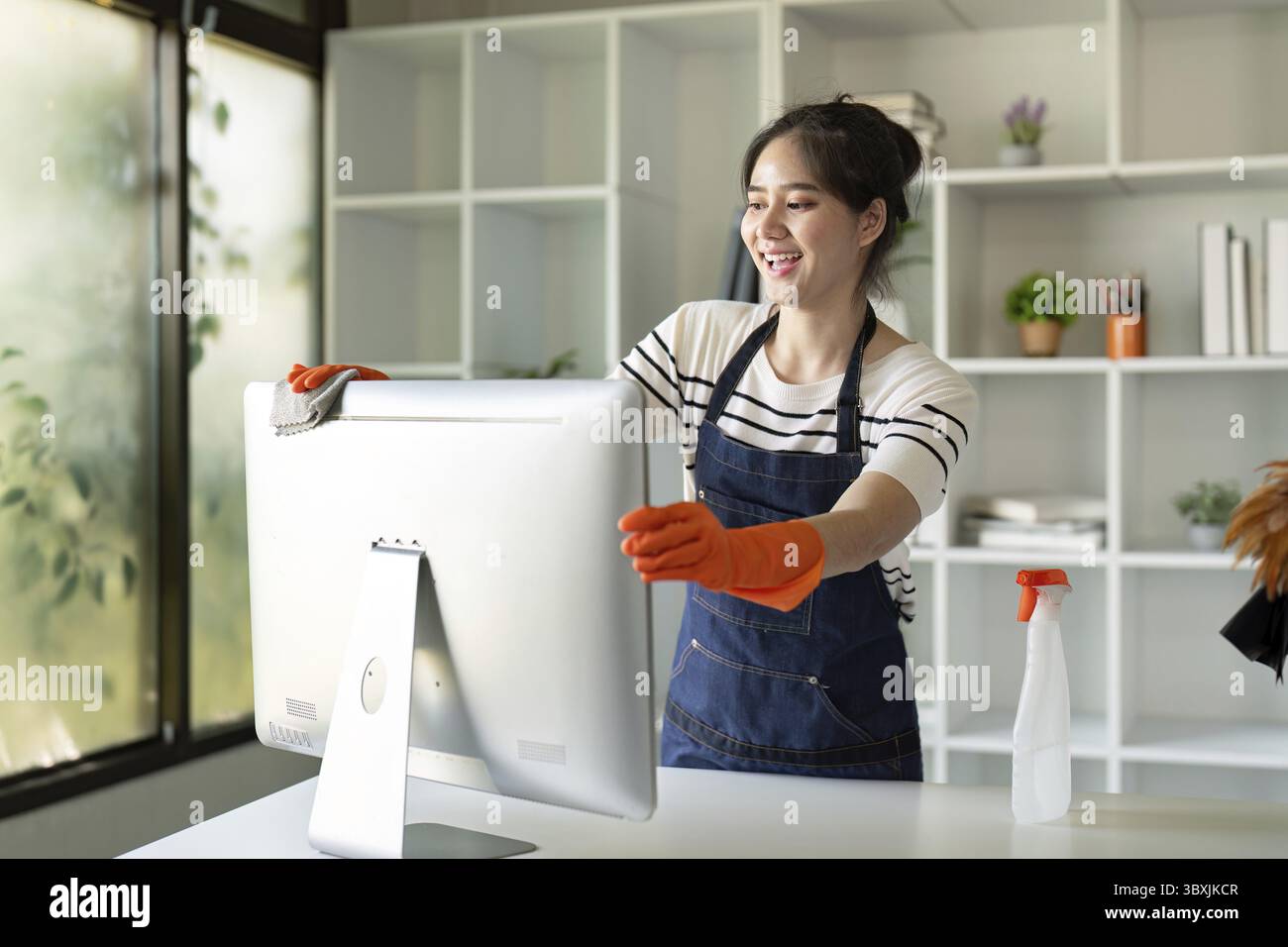 Donna asiatica che indossa guanti che pulisce un monitor in un ufficio domestico, raffigurante un umore gioioso e l'attenzione ai dettagli Foto Stock
