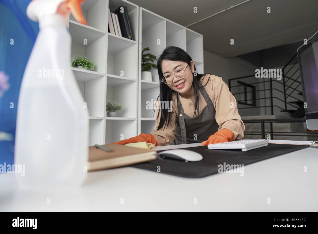Una cameriera allegra che puliva una moderna scrivania con una bottiglia spray e un panno, mostrando un'area di lavoro ordinata e organizzata Foto Stock