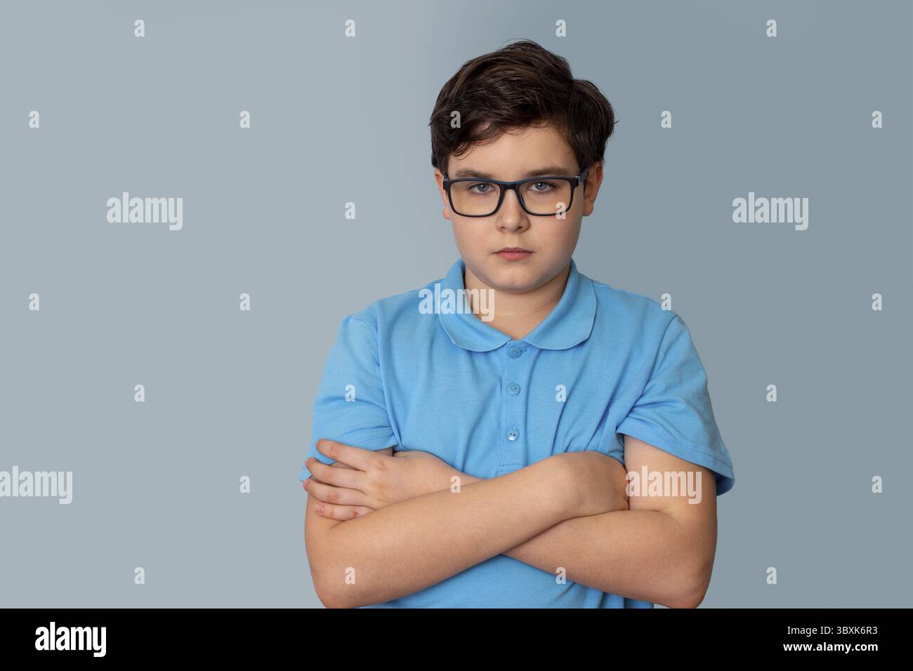 Un ragazzo dai capelli scuri con occhiali, in piedi con una maglietta blu, braccia incrociate sul petto, su sfondo grigio, guarda la fotocamera Foto Stock