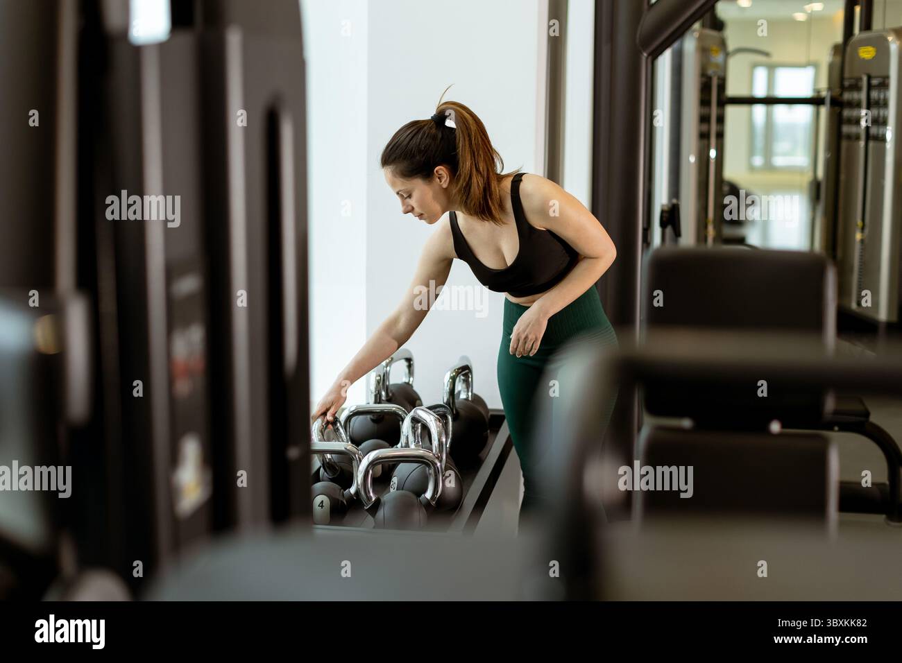 Una donna si concentra sul suo allenamento selezionando i pesi in una palestra contemporanea, coltivando forza e benessere in un ambiente stimolante. Foto Stock