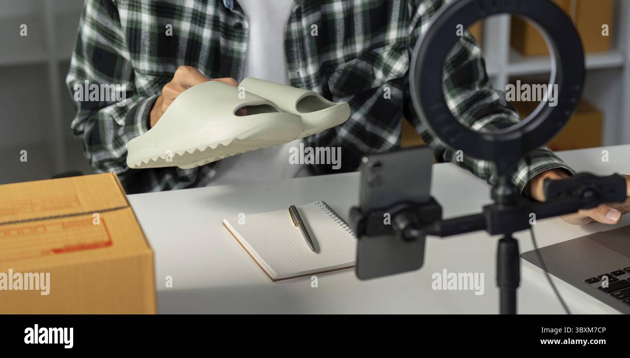 Un giovane imprenditore sta mostrando un prodotto calzaturiero mentre conduce una vendita online, utilizzando uno smartphone e un notebook in uno spazio di lavoro elegante Foto Stock