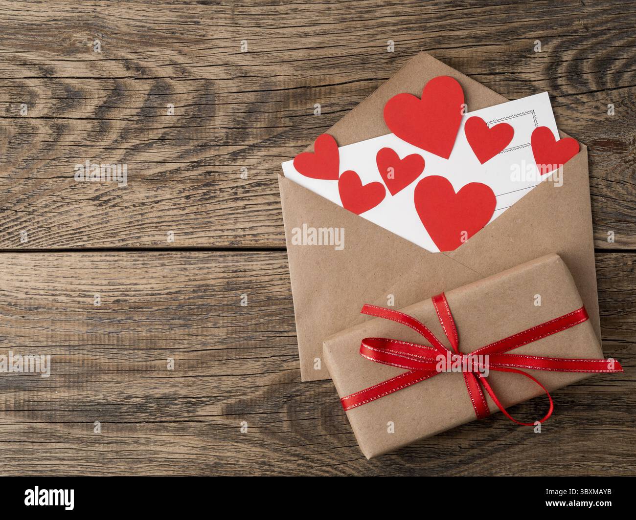 Karte und rote Herzen im offenen Umschlag aus braunem Kraftpapier. Geschenkkasten mit rotem Band auf hoelzernen alten Jahrgang Hintergrund. Gruesse mi Foto Stock