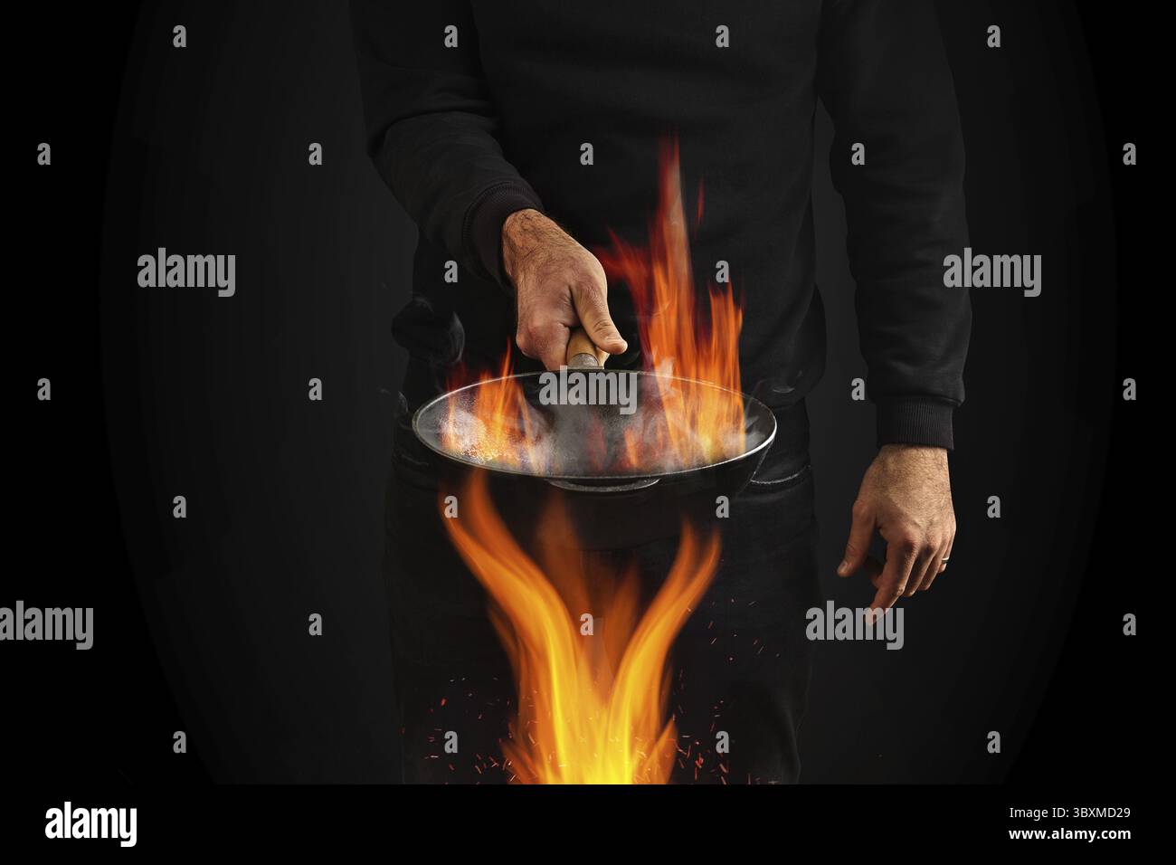 Giovane uomo vestito con jumper e jeans. Sta tenendo il wok pan bruciante con fumo sopra il fuoco, contro sfondo nero studio. Concetto di cucina. Primo piano Foto Stock