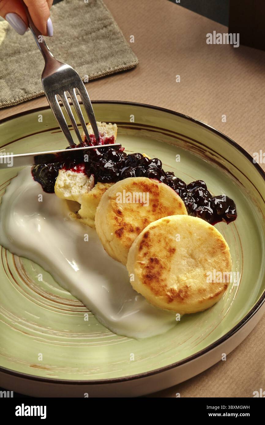 Mani femminili che affettano soffice sciroppo dorato servito su piatto di ceramica con condimento di frutti di bosco e delicata crema di yogurt. Piatto dolce tradizionale ucraino Foto Stock