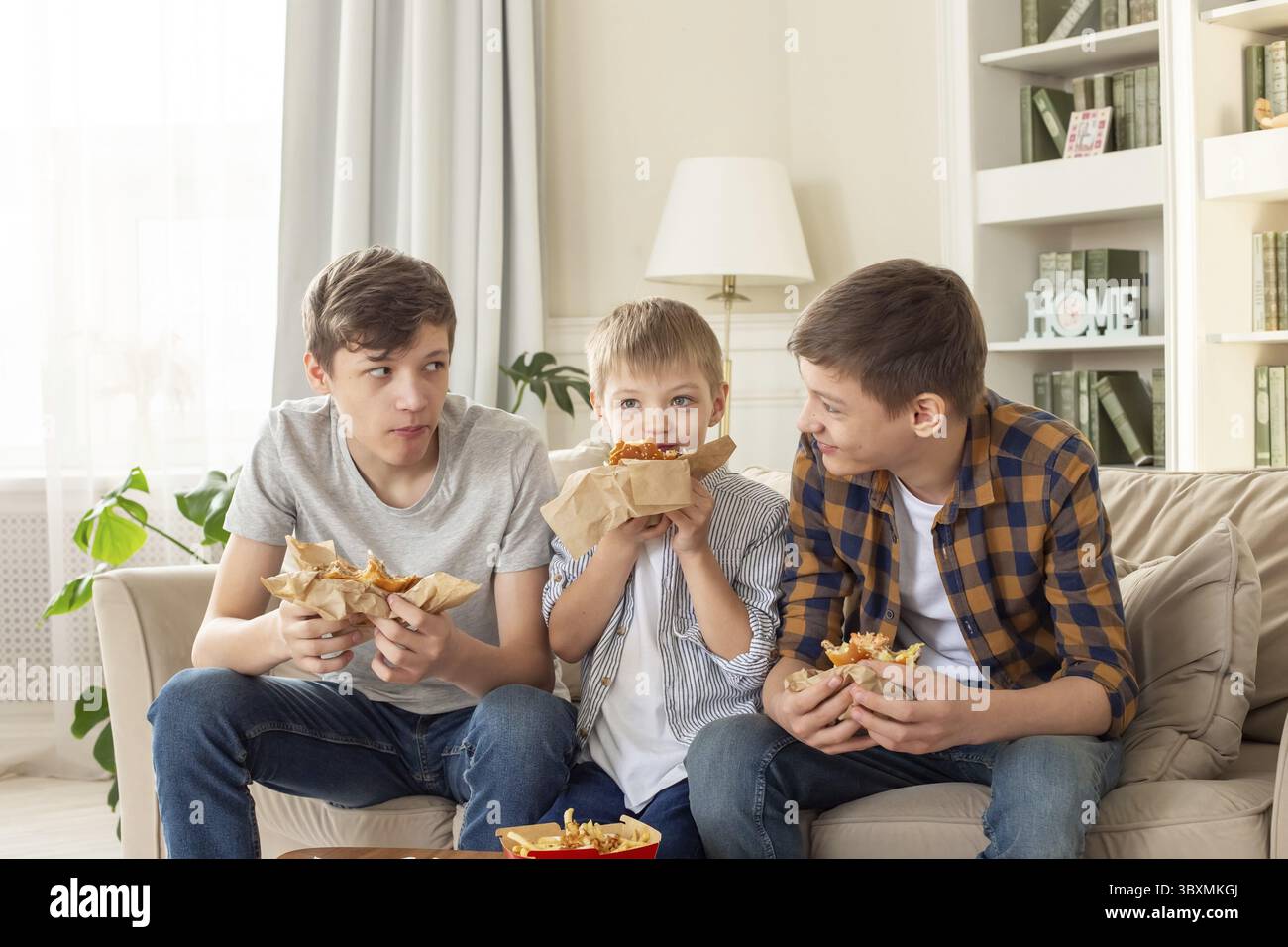 Tre simpatici adolescenti, in abiti casual, si siedono sul divano nel soggiorno, mangiando fast food, un hamburger, comunicazione Foto Stock