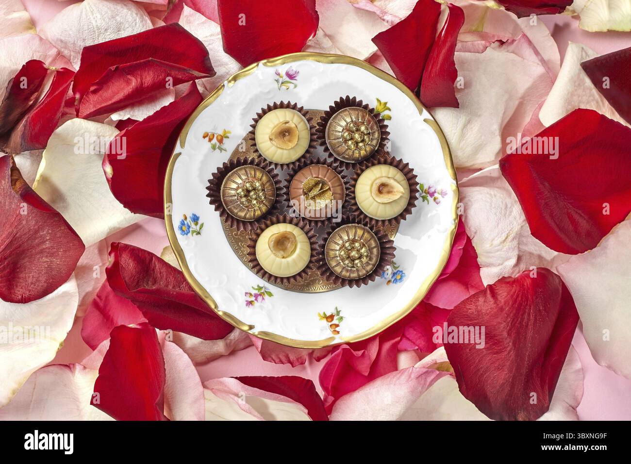 Eleganti cioccolatini artigianali decorati con nocciole, briciole di caramello e perle dorate disposti su piatto vintage con design floreale sullo sfondo di co Foto Stock