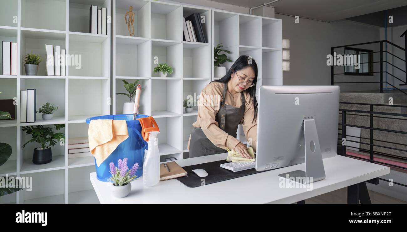 La governante pulisce in uniforme una moderna scrivania con computer e forniture, garantendo uno spazio di lavoro ordinato e organizzato Foto Stock
