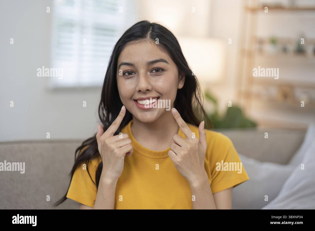 Una giovane donna con una camicia gialla indica con gioia i suoi denti belli e sani mentre sorride con sicurezza in una stanza ben illuminata Foto Stock