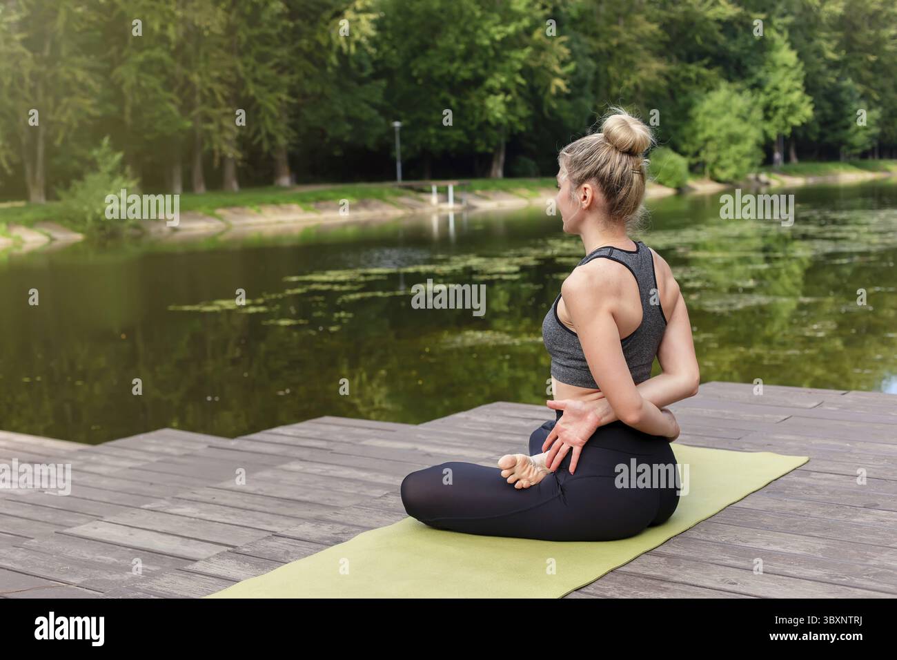 Una donna snella con una parte superiore grigia e leggings, seduta su una piattaforma di legno accanto a uno stagno nel parco in estate e che esegue yoga. Copia spazio Foto Stock