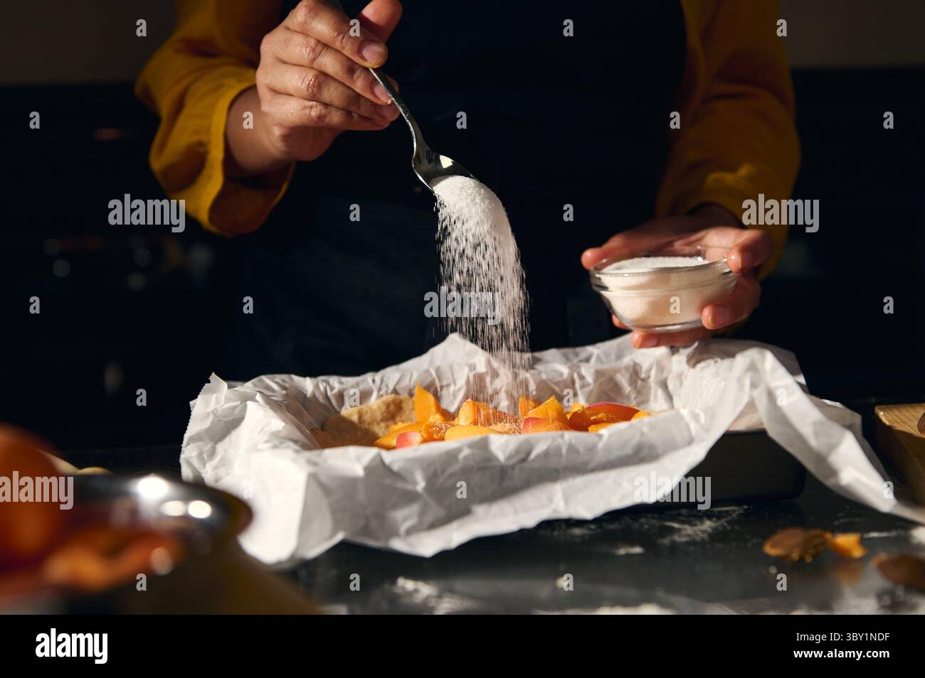 Uno chef prepara una deliziosa ricetta di cottura, spolverando zucchero su ingredienti freschi all'interno di una padella rivestita in pergamena, catturata in un batter d'occhio sotto una luce calda Foto Stock