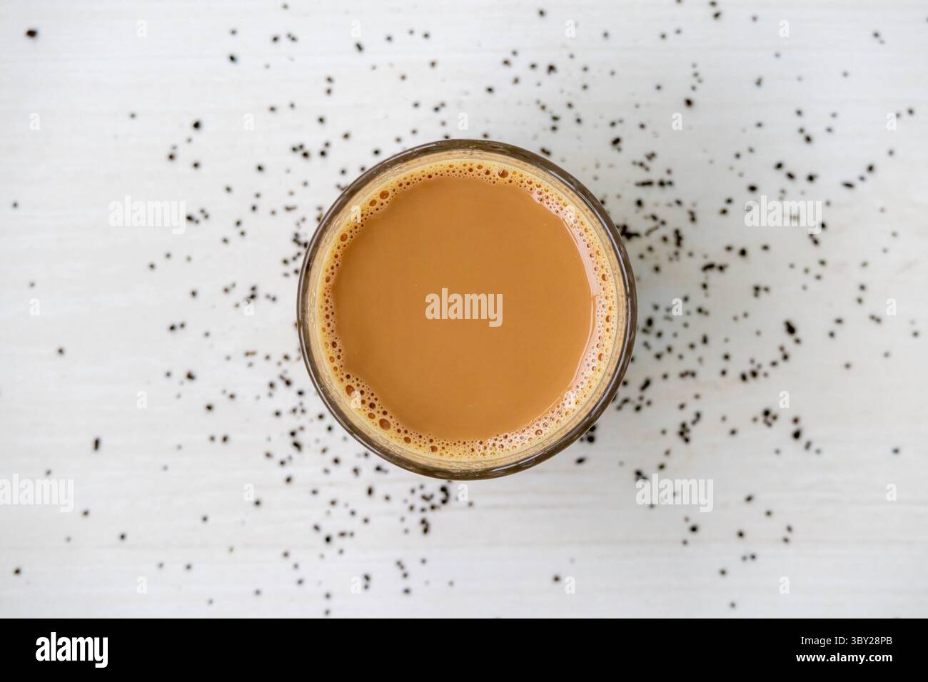 Tè al latte caldo con bollicine in una tazza di vetro, su una superficie di legno chiaro. Granuli di tè sparsi nelle vicinanze. Vista dall'alto della bevanda calda. Foto Stock