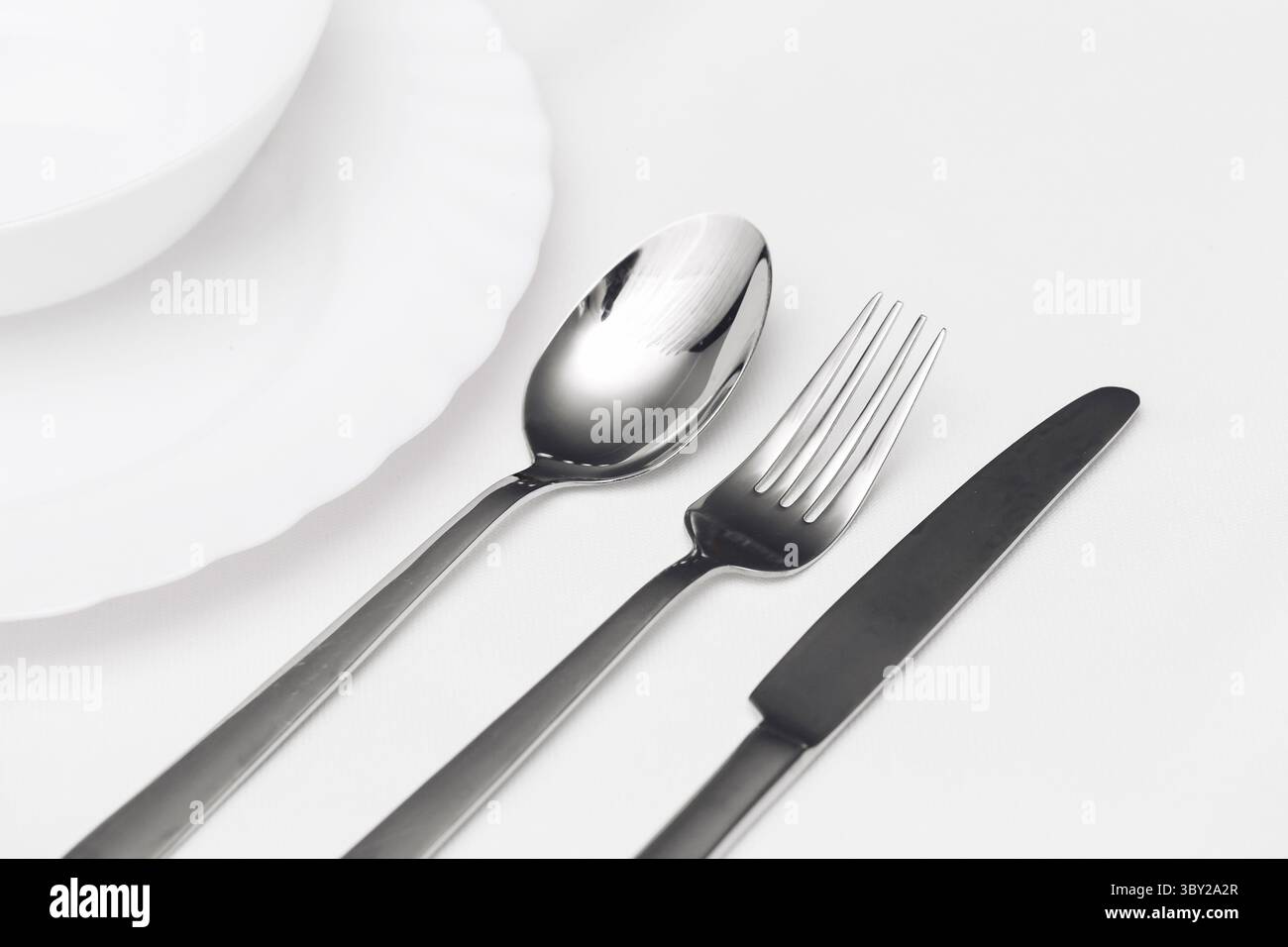 Posate in metallo lucido su sfondo bianco. Coltello, forchetta e cucchiaio accanto alle piastre pulite. Elegante disposizione dei pasti. Utensili in acciaio inox per il pasto. Foto Stock