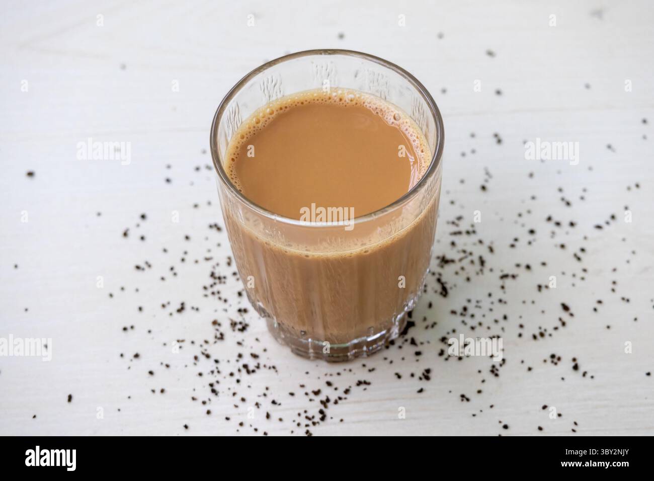 Montare il tè al latte caldo con deliziose bollicine, servito in una tazza di vetro trasparente su un tavolo di legno chiaro. Foglie di tè sciolte circondano elegantemente la tazza. Foto Stock