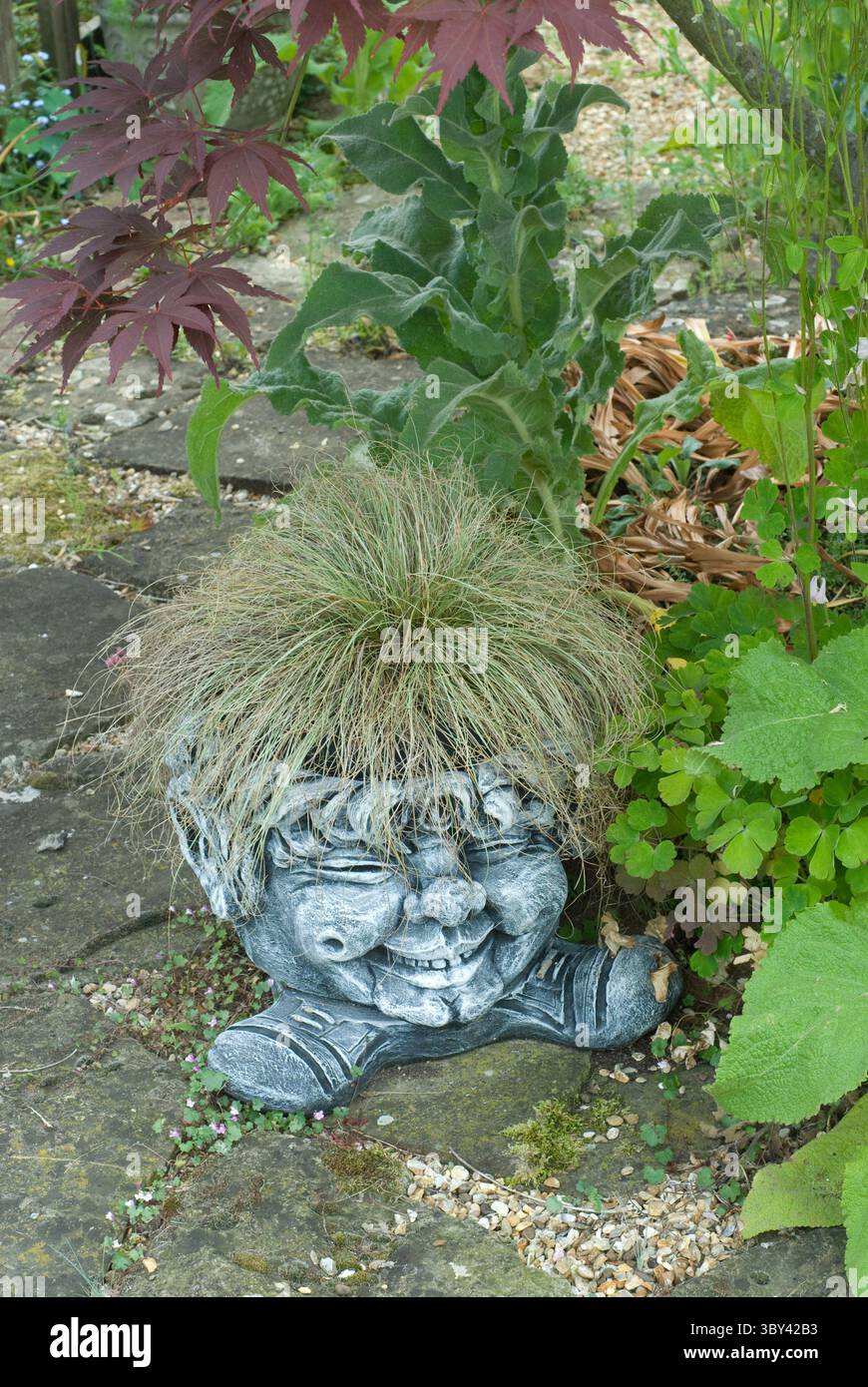 Una scultura stravagante che assomiglia a una figura sorridente adornata di erba come capelli, circondata da lussureggianti piante verdi in un giardino sereno. Foto Stock