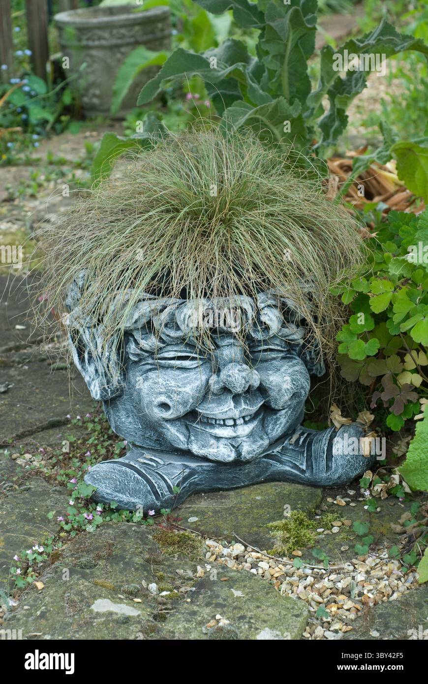 Una stravagante scultura in pietra con un'espressione gioiosa sfoggia un ciuffo di erba lussureggiante come i suoi capelli, aggiungendo fascino all'ambiente del giardino. Foto Stock