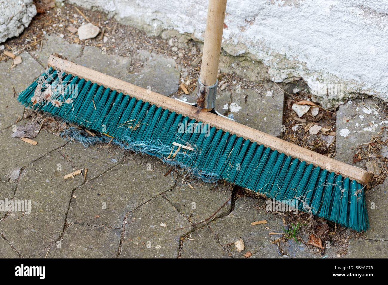 Primo piano di una scopa a pressione color ottanio con una maniglia in legno appoggiata su un patio in cemento incrinato accanto a un muro di cemento grezzo. Foto Stock