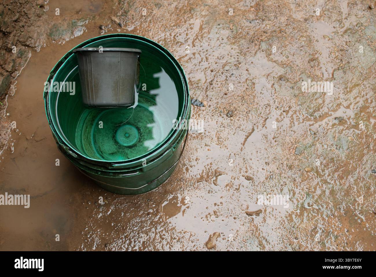 Un secchio verde pieno d'acqua e una tazza di metallo poggiano su un terreno fangoso, riflettendo le dure condizioni della raccolta dell'acqua nelle zone rurali e della vita quotidiana. Foto Stock