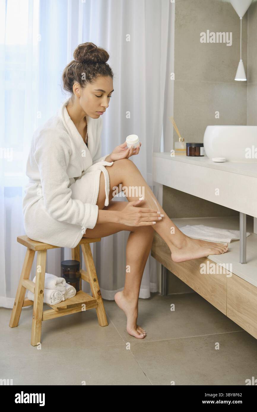 Una donna siede su uno sgabello in un bagno, concentrandosi sull'applicazione della lozione alla gamba. La luce naturale si diffonde, mettendo in evidenza la sua dedizione alla cura della persona Foto Stock