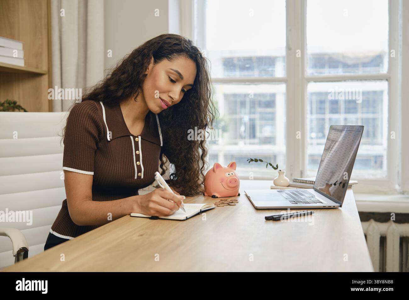 Una donna siede su una scrivania di legno, scrive su un notebook e pianifica le proprie finanze in un luminoso ufficio domestico. Un portatile e un salvadanaio rosa sono visibili, su Foto Stock
