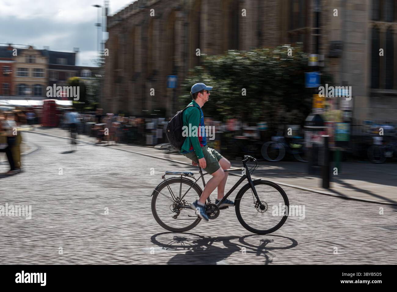 Cambridge , Inghilterra, Regno Unito - 13 luglio 2025: Panoramica di una foto di un ciclista che cavalca a mani libere attraverso la soleggiata strada della città nel campus con movimento sfocato backgroun Foto Stock