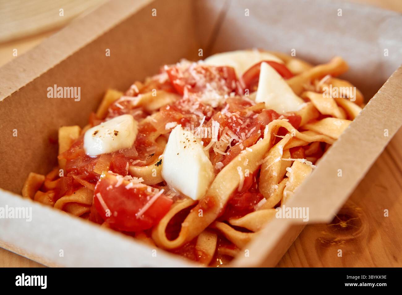 Tagliatelle fresche servite in scatola di cartone, condite con mozzarella, pomodori succosi ed erbe grattugiate per un delizioso piatto italiano. Foto Stock