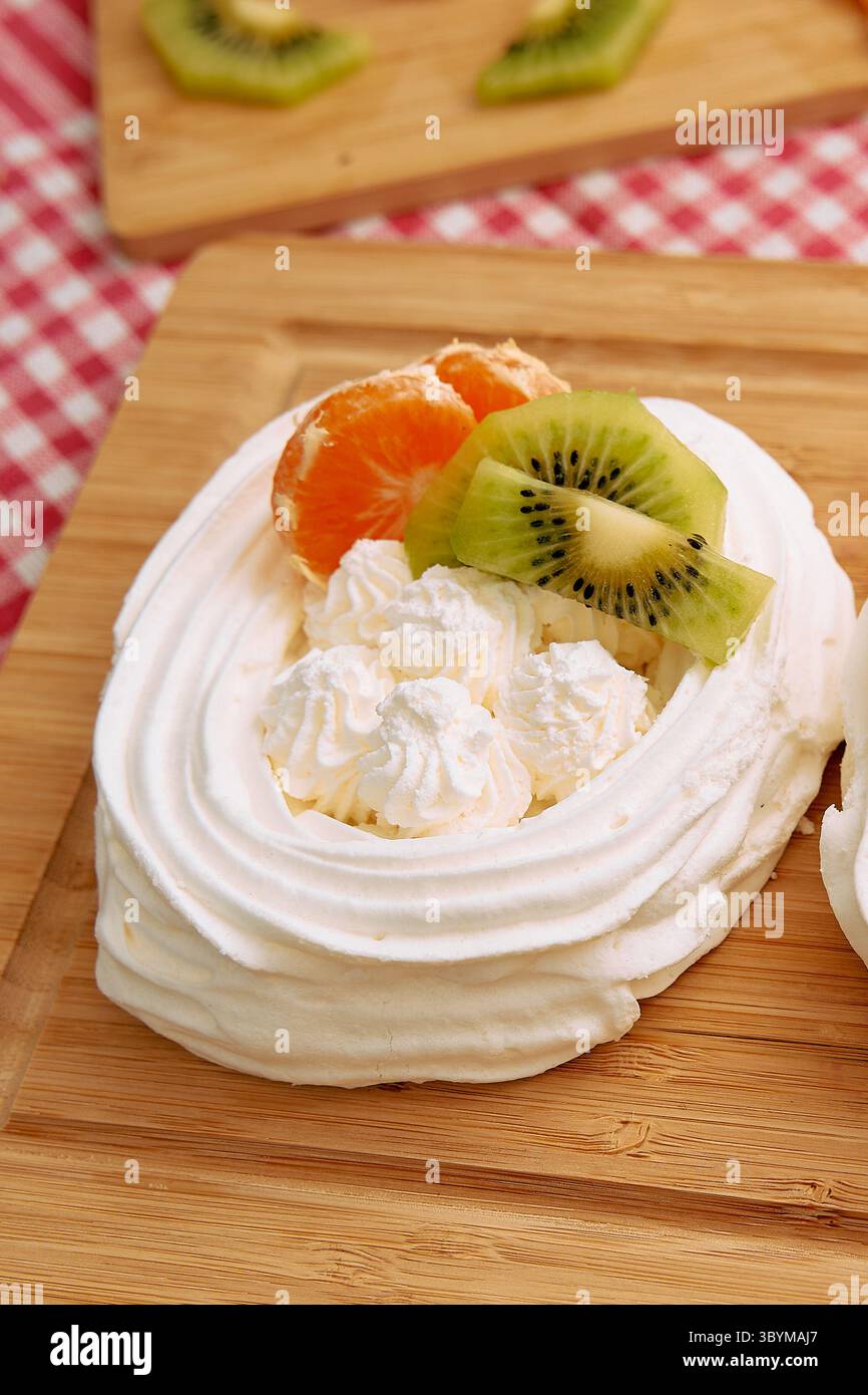Primo piano di un dessert pavlova adornato con panna montata, fette di mandarino e kiwi su una tavola di legno, mostrando una miscela perfetta di soffici, dolci, Foto Stock