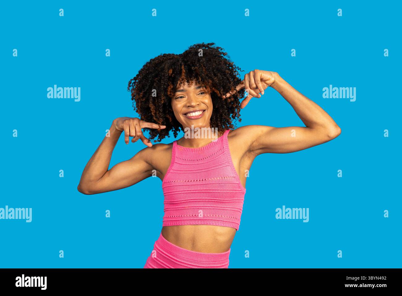 Donna sportiva sorridente che punta i bicipiti promuovendo al contempo la cura di sé e la positività del corpo Foto Stock