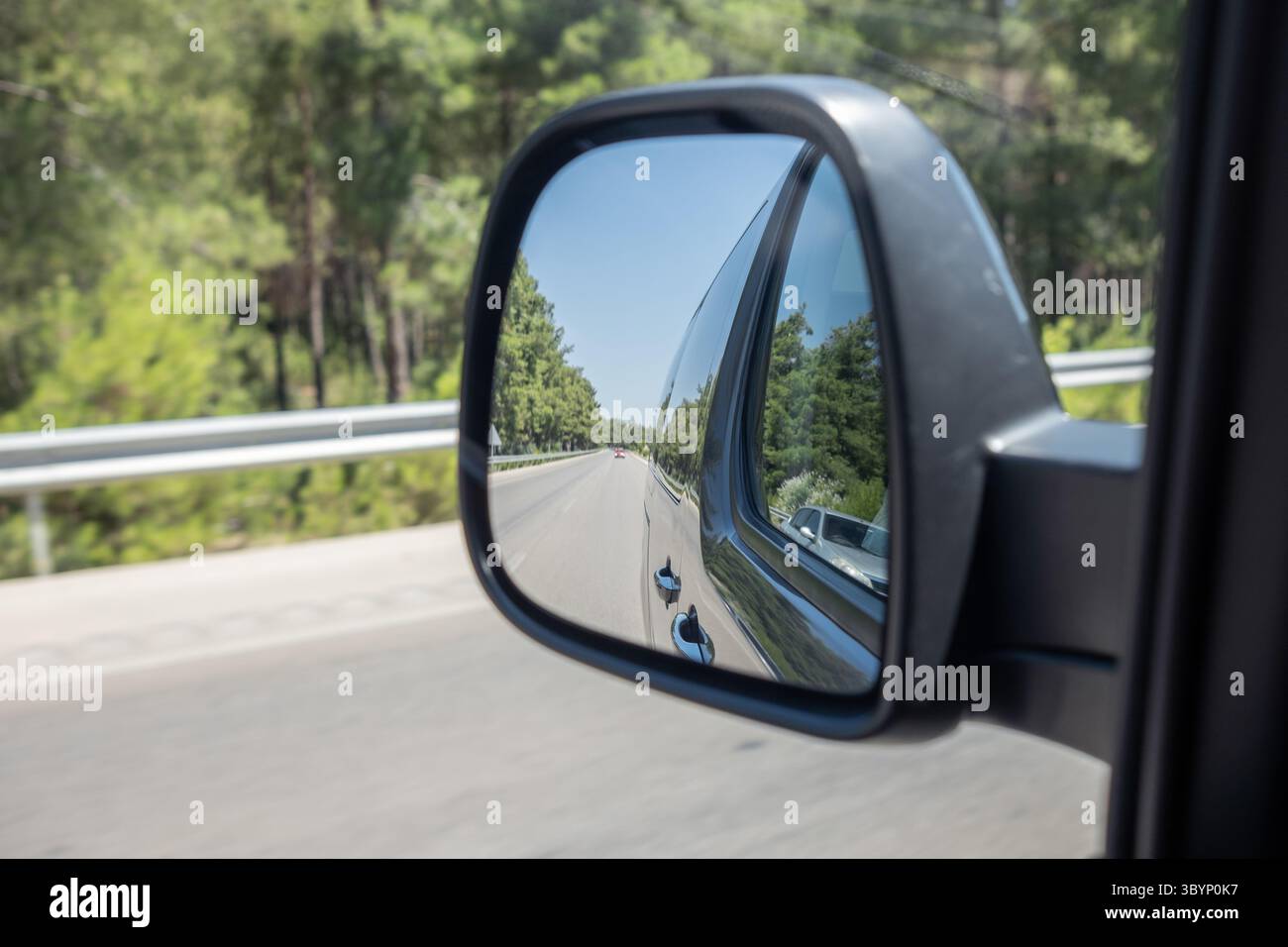 Uno specchio laterale cattura la vista di una strada tortuosa costeggiata da alberi verdi sotto un cielo azzurro limpido durante una giornata di sole, evidenziando il viaggio da percorrere. Foto Stock