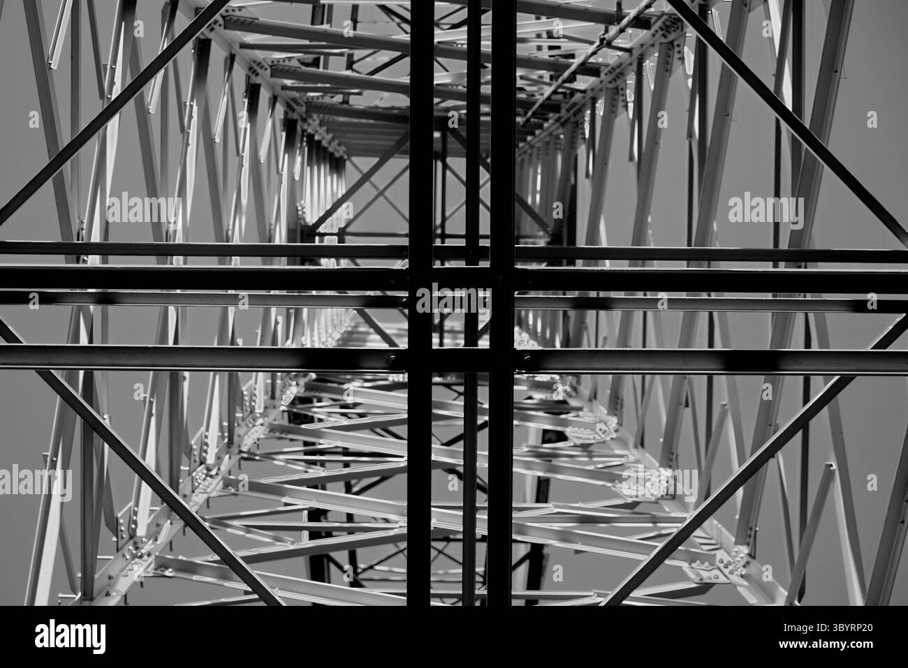 Guardando verso l'alto attraverso l'intricata struttura in acciaio di una torre di trasmissione ad alta tensione posizionata contro un cielo limpido, rivelando audaci motivi industriali. Foto Stock
