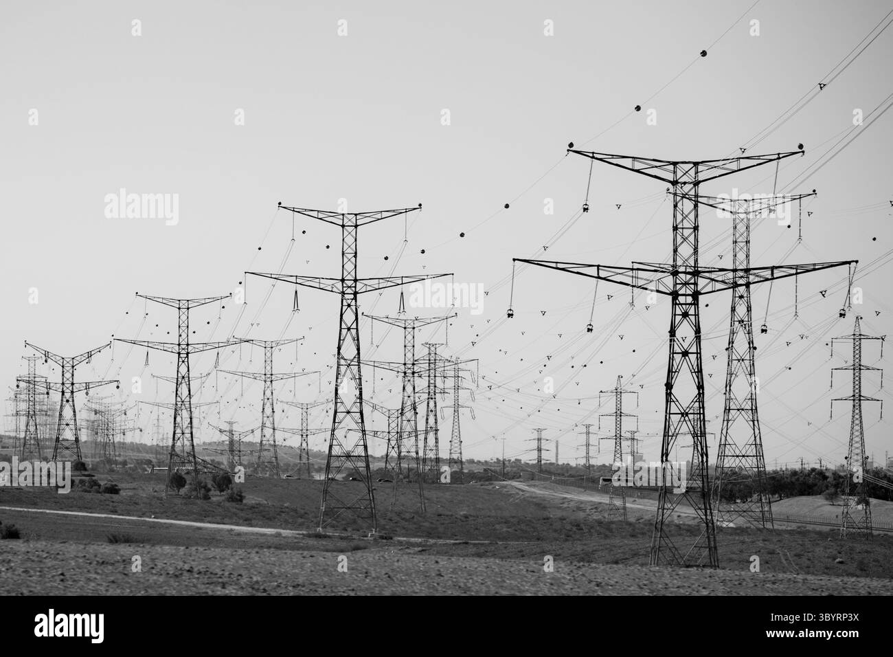 Le torri di trasmissione ad alta tensione illuminate dal sole si estendono attraverso un orizzonte rurale sotto un cielo azzurro, evidenziando scala industriale e linee geometriche audaci. Foto Stock