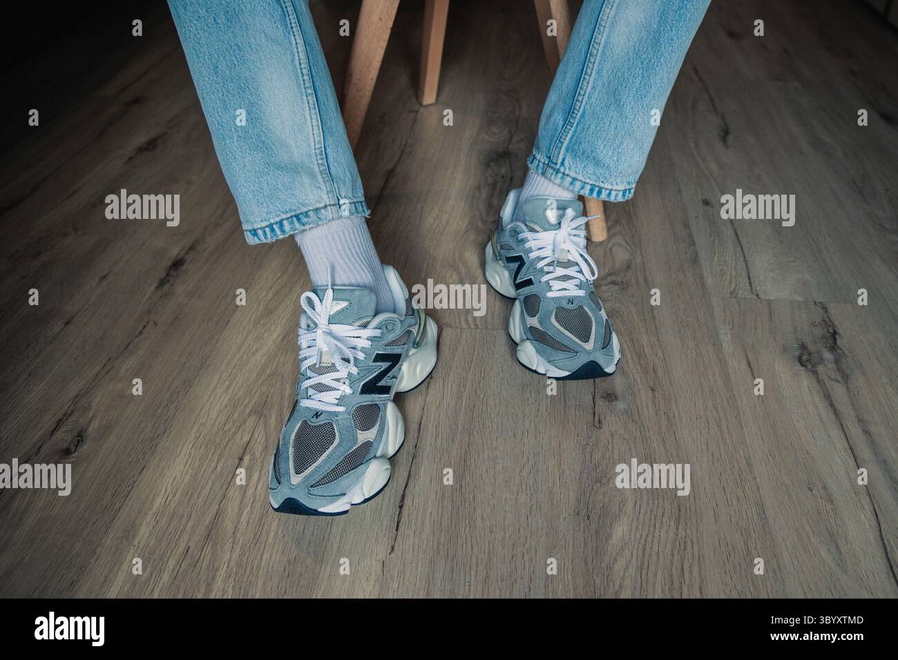 Vista frontale della persona che indossa scarpe da ginnastica New Balance 9060 con calze bianche e jeans azzurri seduti su pavimento in legno, sottolineando lo streetwear casual Foto Stock