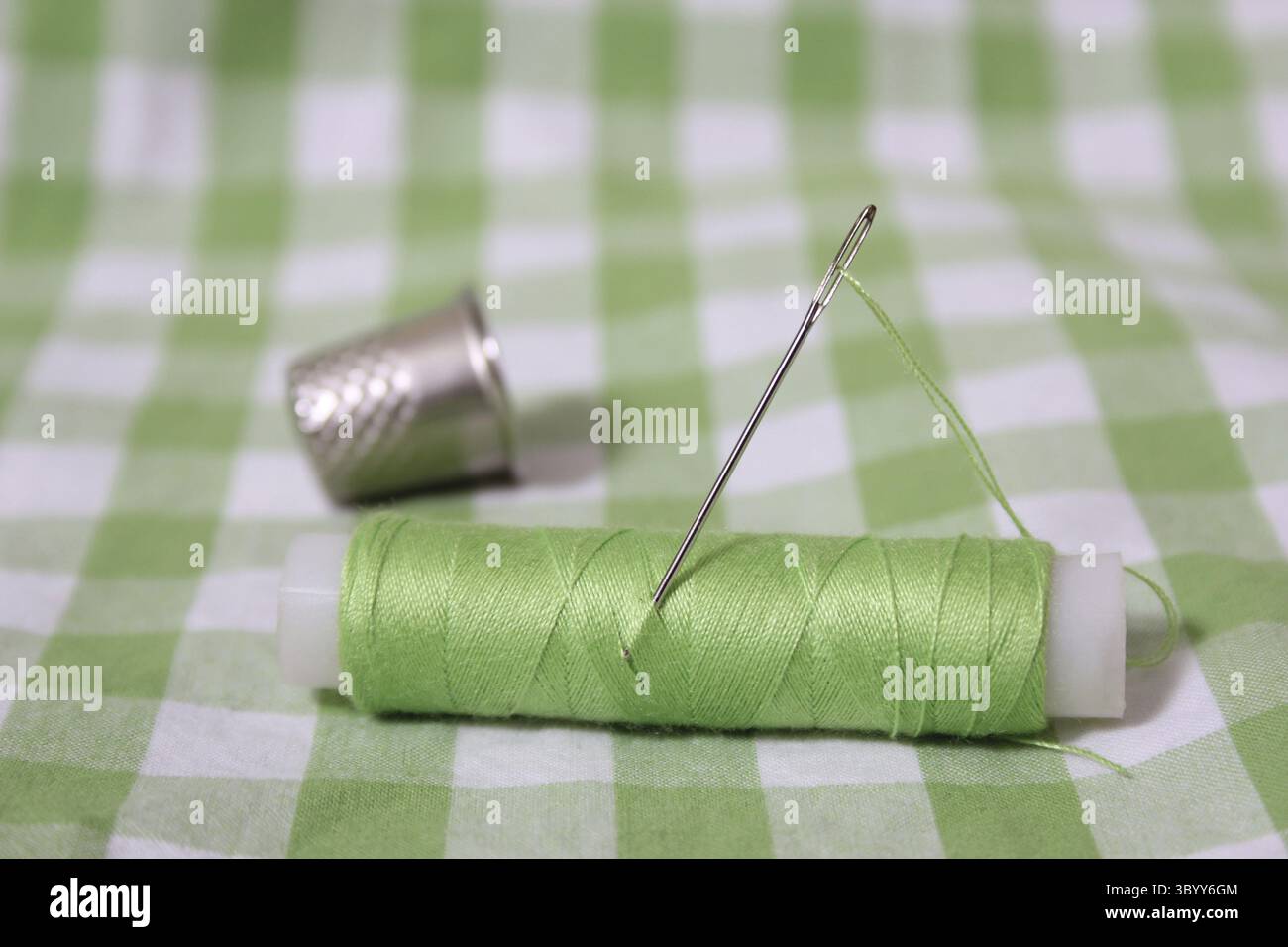 Bobina con filettatura verde chiaro, ditale e ago su tessuto a scacchi verde Foto Stock