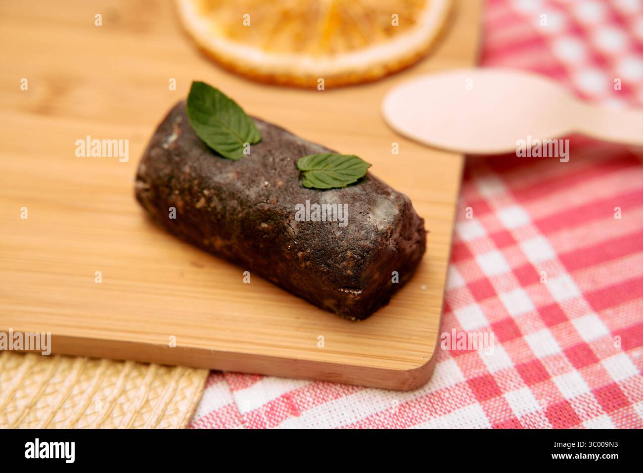 Un ricco tronco di ganache al cioccolato adornato con foglie di menta si trova su una tavola di legno, posta su un panno di gingham rosso. nelle vicinanze si trovano un cucchiaio di legno e un secco Foto Stock
