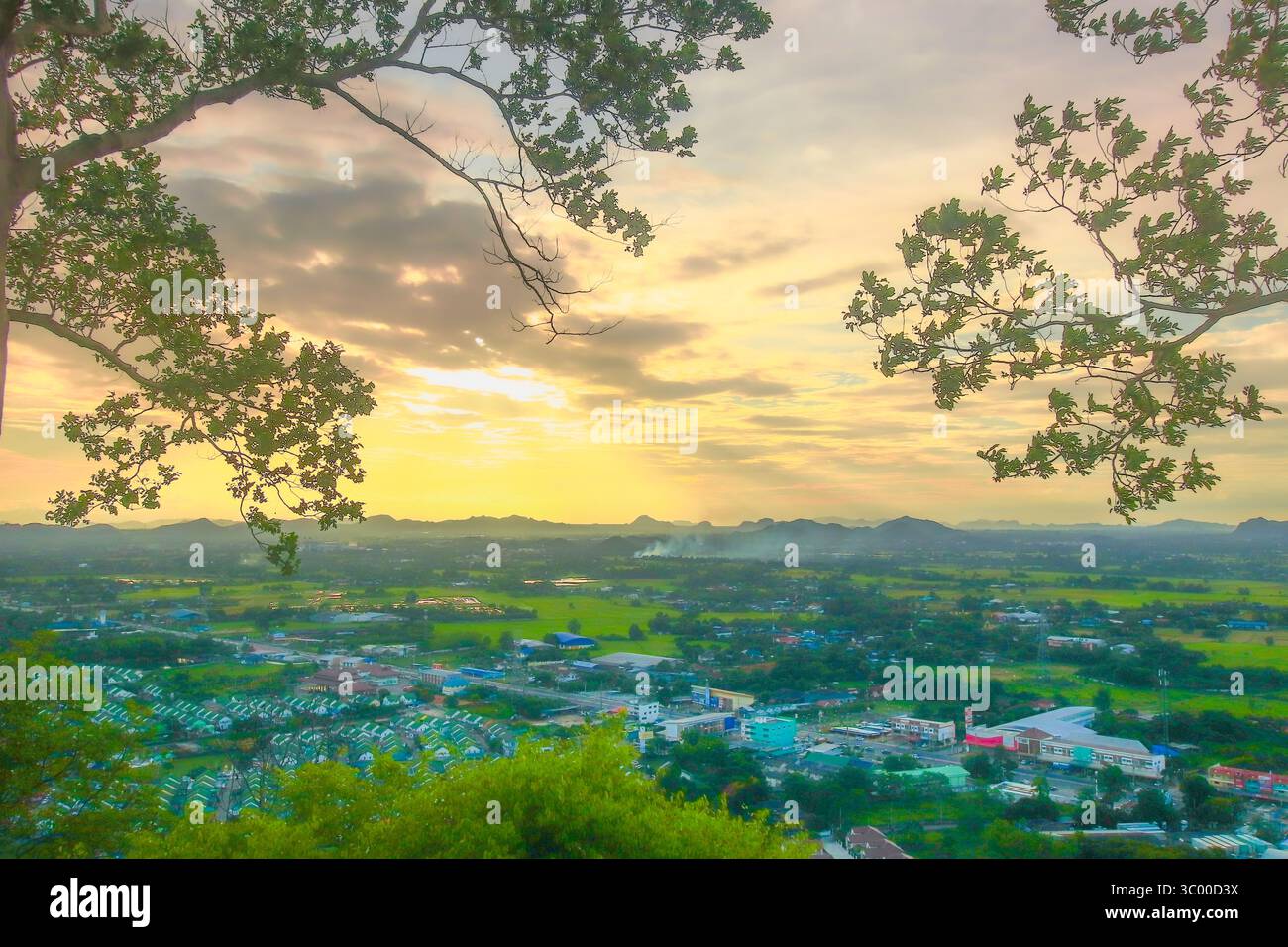 Vista panoramica al tramonto dorato di una cittadina di campagna tailandese, incorniciata da rami di alberi con campi verdi, case e montagne lontane. Foto Stock