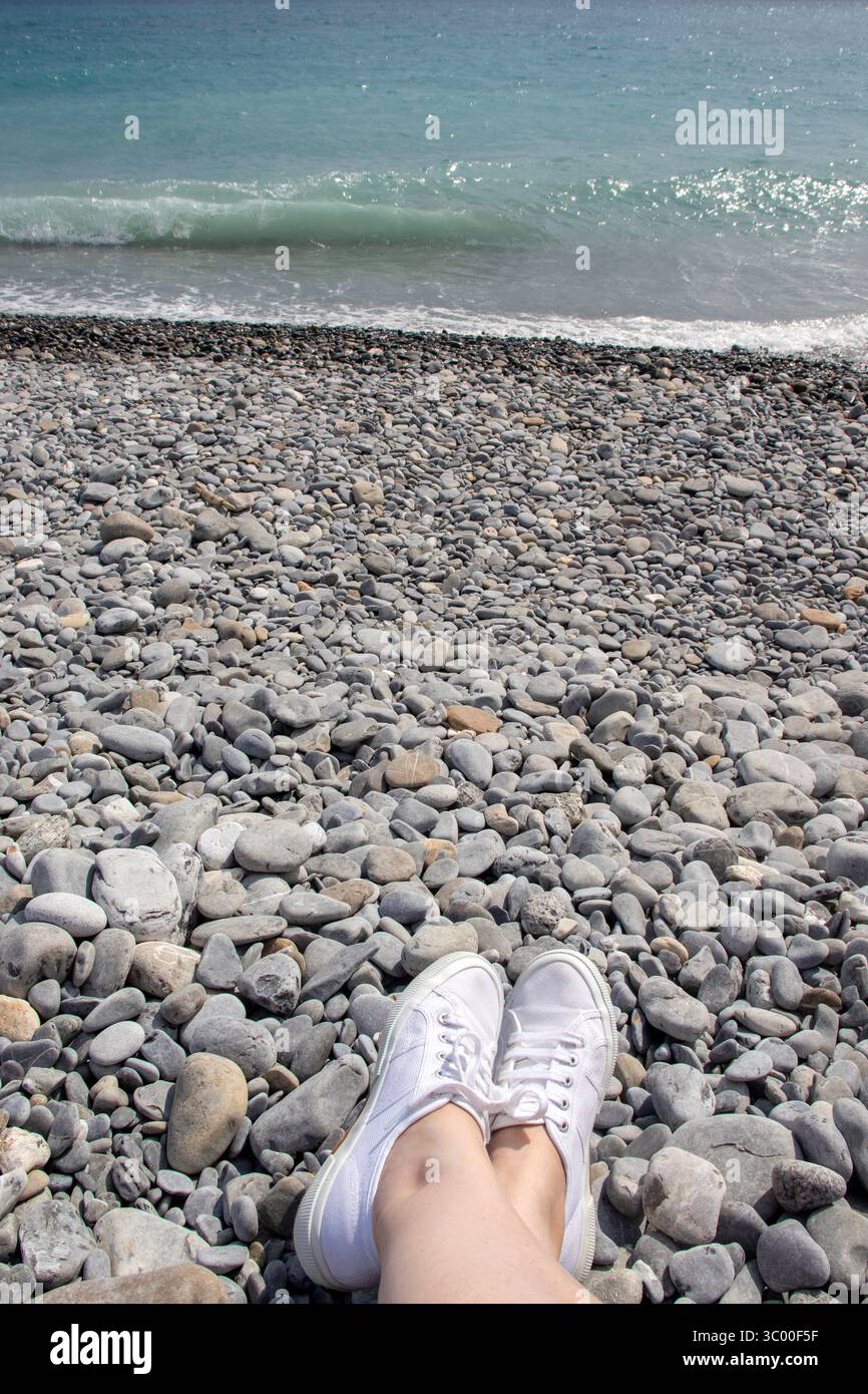 Nessun problema! I piedi casual indossati con scarpe bianche in tela estiva sono rilassati in una posizione incrociata alla caviglia su una spiaggia italiana di ciottoli grigi sul Mediterraneo Foto Stock