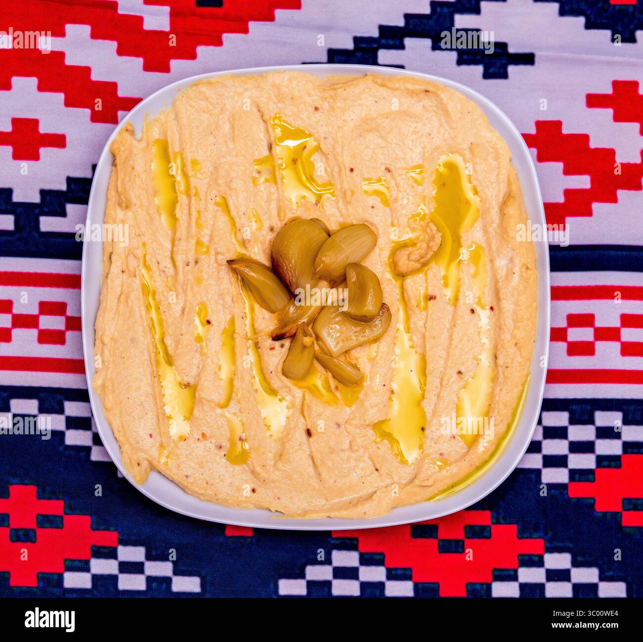 Hummus cremoso, coronato da spicchi d'aglio arrostiti e nastri d'olio d'oliva dorati su un vivace tessuto geometrico Foto Stock